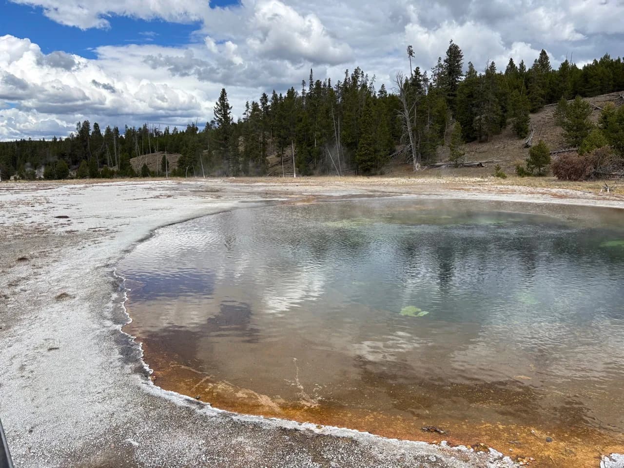 Reflective geothermal pool bordered by mineral ground and forest.